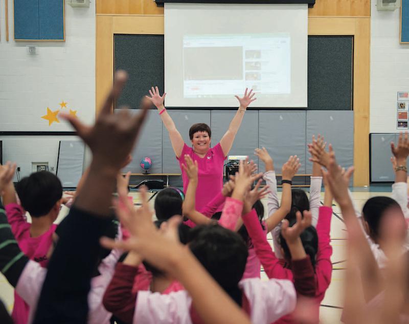 students in gymnasium mimicking teacher who has her hands in the air