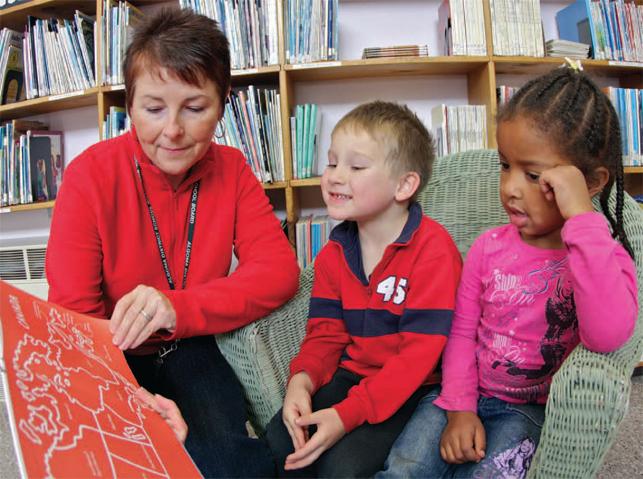 teacher holding a picture book reading to two students in a library