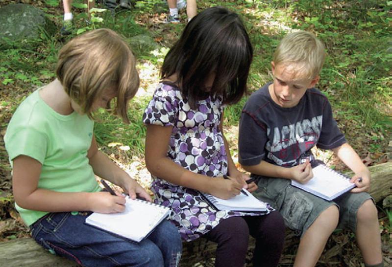 three young elementary students sitting on stone outside writing in notebooks
