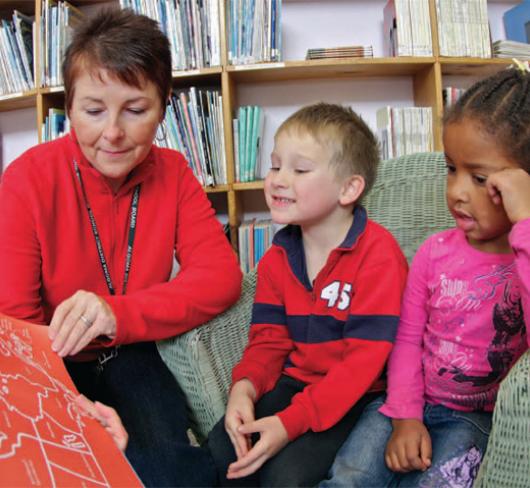teacher holding a picture book reading to two students in a library