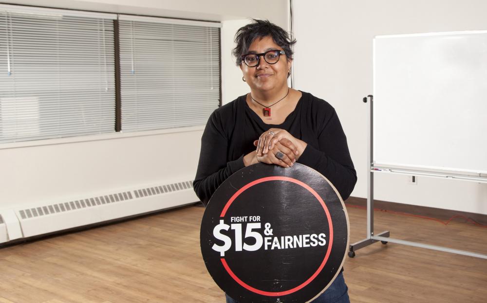 Deena Ladd standing in class holding $15 & Fairness logo