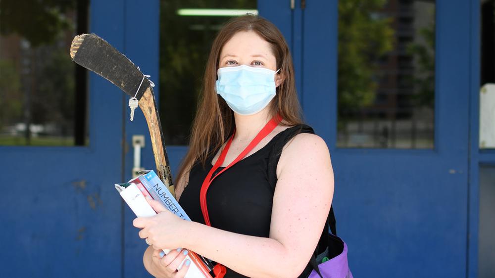 Courtney White standing in front of school wearing mask, carrying books and hockey stick