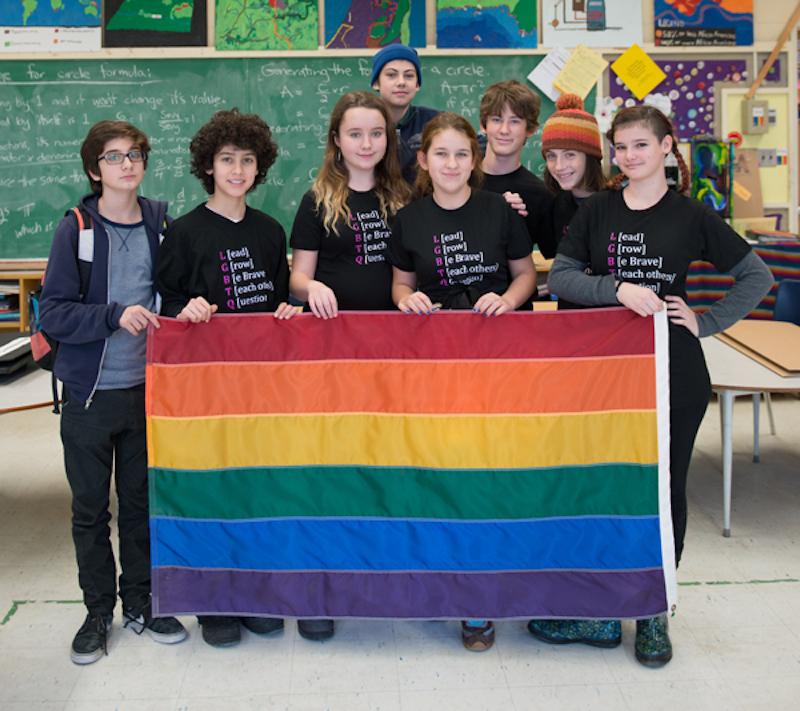 Students standing in classroom holding a rainbow flag
