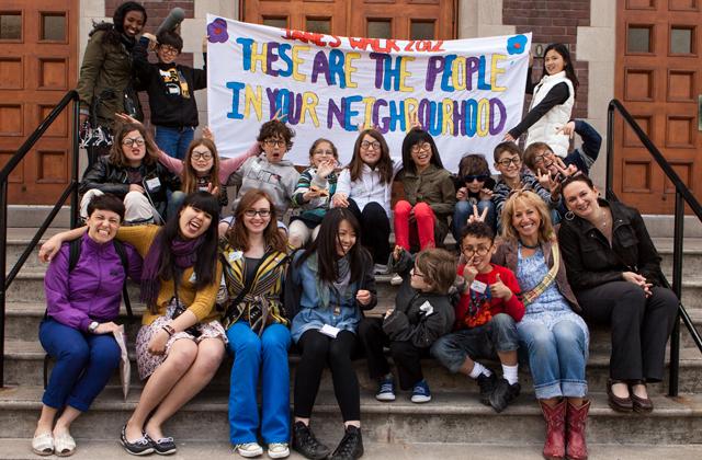 students sitting together holding sign that reads "These are people in our neighbourhood"