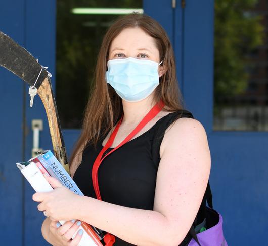 Courtney White standing in front of school wearing mask, carrying books and hockey stick