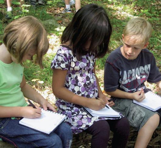 three young elementary students sitting on stone outside writing in notebooks