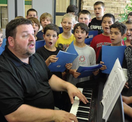 music teacher singing and playing keyboard surrounded by students who are also singing