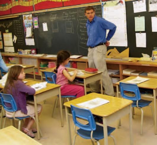 teacher standing in front of classroom while children are working
